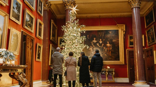 Four visitors stand beside a large illuminated Christmas tree in Attingham Park's Picture Gallery. The tree is decorated with sparkling ornaments and topped with a star, surrounded by red walls, grand columns and framed paintings, including a large family portrait hanging behind the tree.